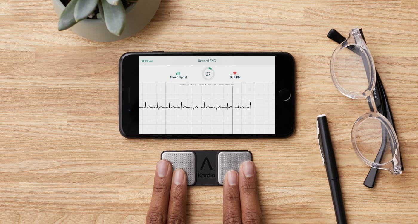 fingers on the KardiaMobile electrode next to a phone showing the single lead EKG on a desk with a pencil and glasses